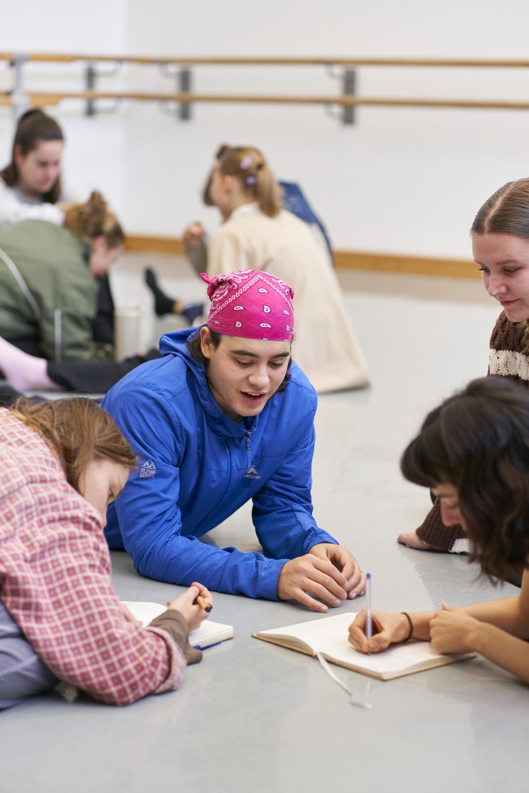 A group of students sitting and lying in a circle writing notes in a rehearsal room